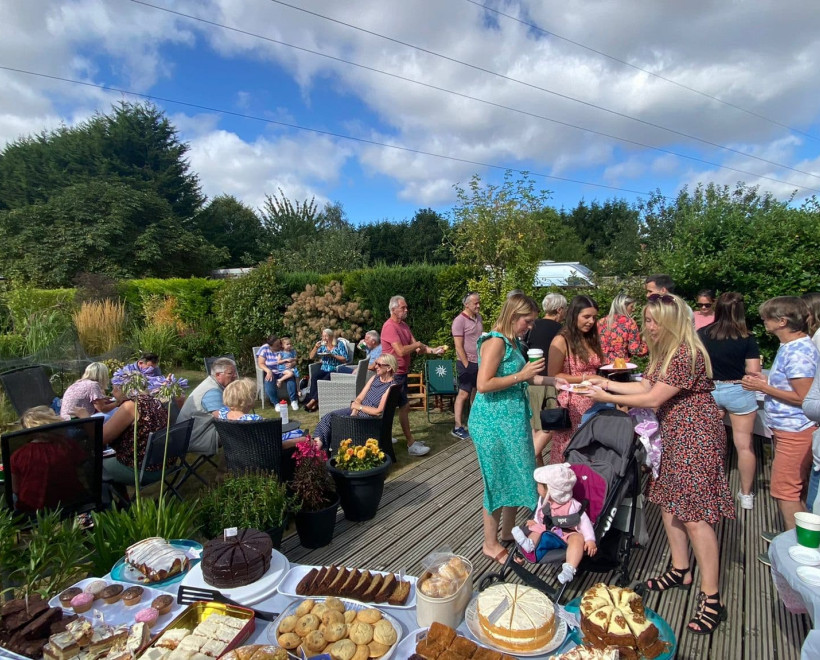 Dozens of people in garden with hedges, wooden decking and blue sky. There is a table full of cakes and baked goods in the foreground with people standing and sitting.