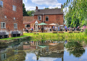 Red brick historic building the Watermill in Middle Rasen, with people sitting on benches with parasols. In the foreground is a body of water with greenery.
