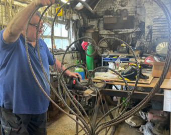 A man, Stefanos, a blacksmith who has supported St Barnabas in the past. Stood in his workshop wearing blue polo shirt and working on large metal heart sculpture.