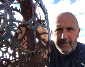 Kevin Baumber, one of the Artists for the St Barnabas HeART Trail. Man with beard posing with sculpture depicting industrial cogs made of metal.