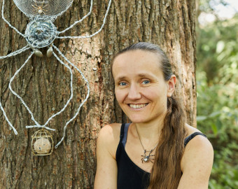 Lois Cordelia, one of the Artists for the St Barnabas HeART Trail. Woman with brown hair in ponytail by large tree trunk with silver spider sculpture made of twisted wires.