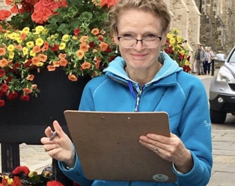 Karin Christensen, one of the Artists for the St Barnabas HeART Trail. Woman with short blonde hair, wearing blue fleece jacket and holding clipboard by colourful flowers