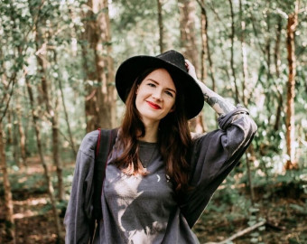 Rosie Ablewhite, one of the Artists for the St Barnabas HeART Trail. Woman with long brown hair wearing black hat and top, smiling with hand on head. In background there are trees.