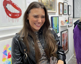 Sian Bristow, one of the Artists for the St Barnabas HeART Trail. Woman with long brown hair wearing leather jacket, smiling with paintings on wall behind her.