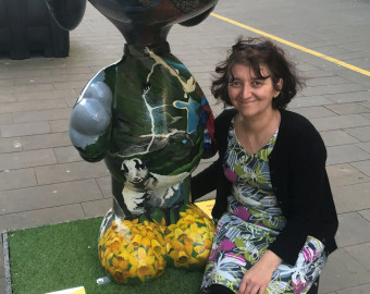 Kathleen Smith, one of the Artists for the St Barnabas HeART Trail. Woman with short black hair, wearing black and tropical print clothing, sitting by a statue painted with wildlife theme.