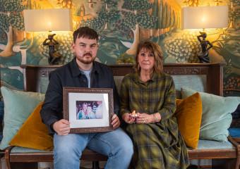 A young man and a women, sat on a sofa, within their home. Being held by the man is a picture of a woman, his mother.