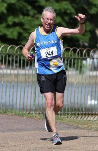 Man running in blue St Barnabas Hospice top with runner number, black shorts, with one arm raised