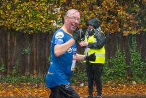 A man running in the rain, wearing a blue St Barnabas Hospice running shirt, and giving the thumbs up