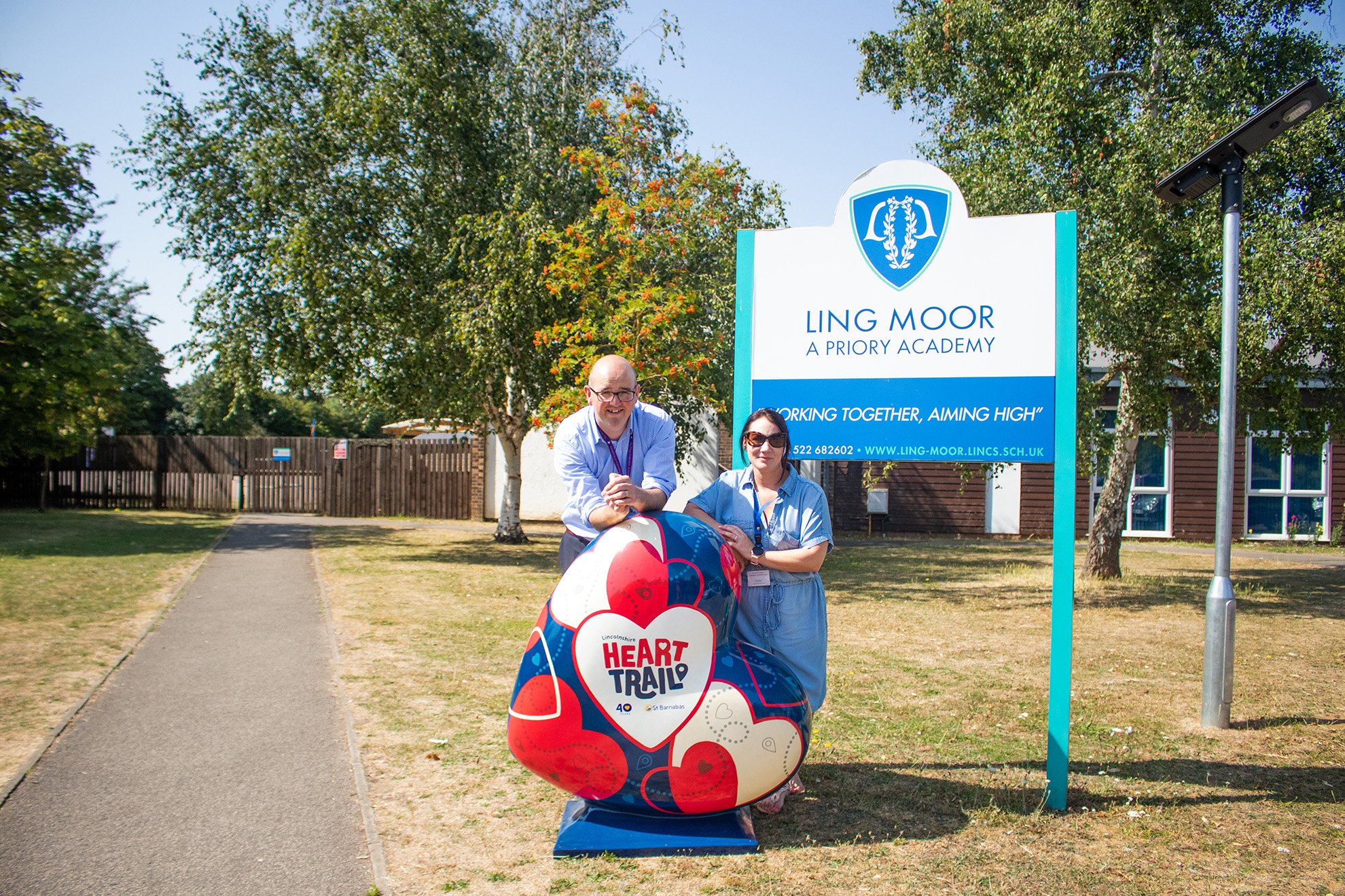 Two people. Simon, the headteacher of Lingmoor Primary Academy and Veronica, Head of Fundraising at St Barnabas Hospice. Both are stood behind the HeART Trail flasghsip hear, outside Lingmoor Primary