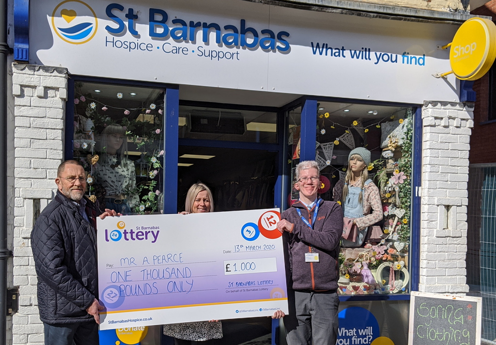 Two men and a woman with a large presenting cheque in front of St Barnabas Hospice charity shop.