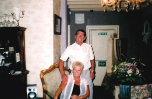 Jean and Keith Bray, a man standing up and a woman sitting on a chair, both are smiling at the camera.