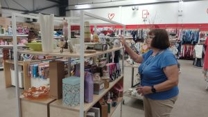 Woman with short brown hair and glasses, wearing blue T-shirt, putting items on shelf in St Barnabas Hospice charity shop