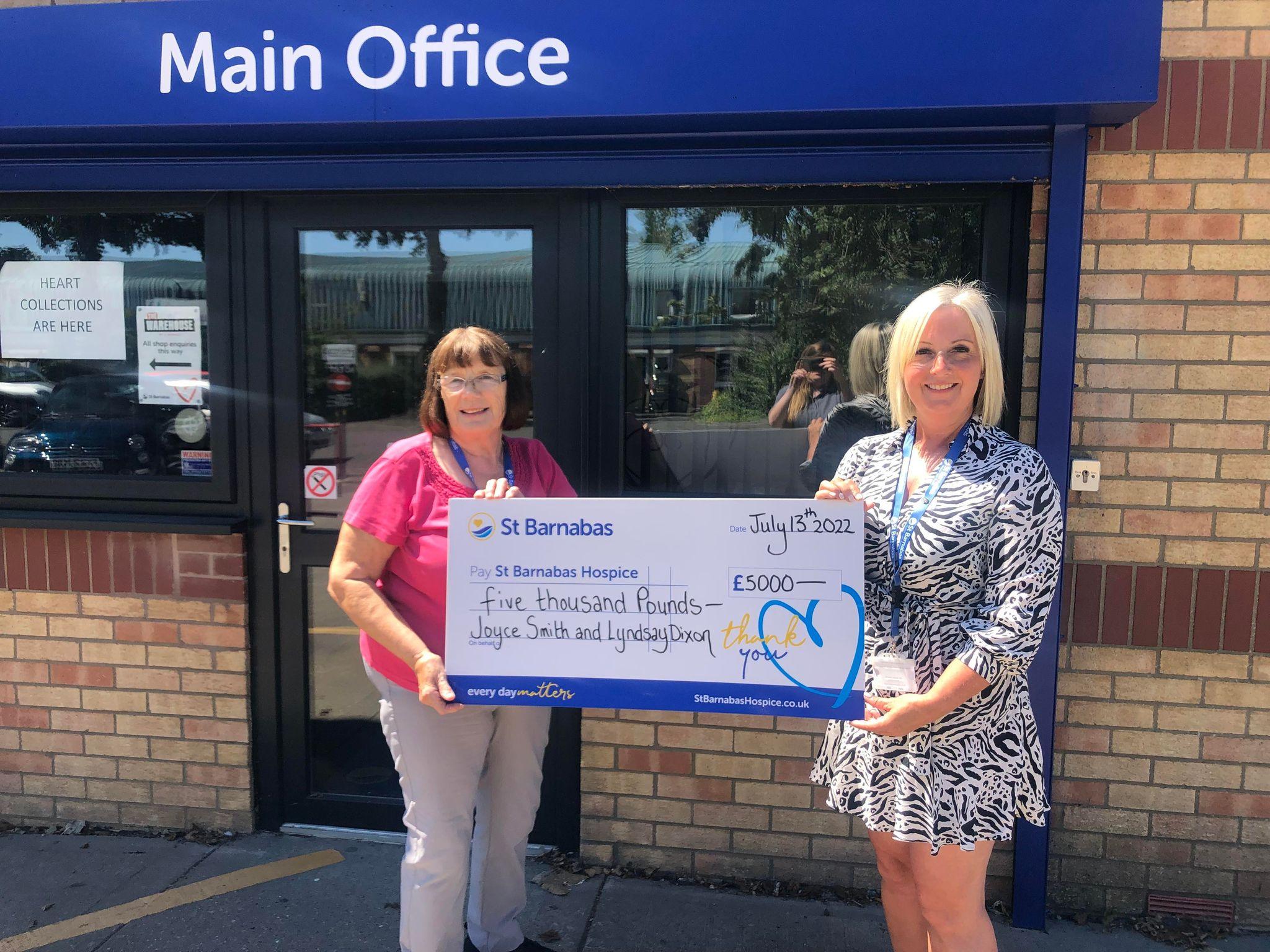 Two women holding a large presentation cheque in front of blue sign with white text "Main Office"