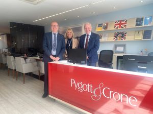 Two men and blonde woman standing at red Pygott & Crone branded desk