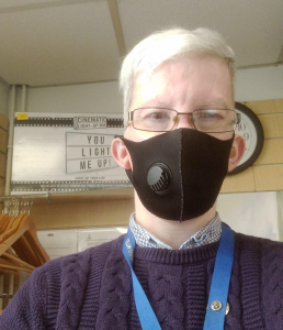 Man with silver hair and glasses wearing a black facemask, dark blue jumper, in a shop environment.