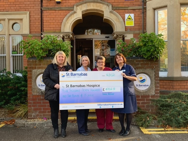 Four women holding large white and blue presentation cheque in front of the red brick Inpatient Unit in Lincoln,