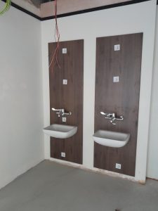 Two white sinks at accessible height with chrome taps against wooden paneling on white wall.