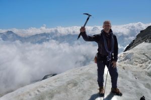 Man holding pick and wearing safety harness on top of mountain summit, surrounded by clouds