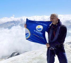 Man holding blue St Barnabas Hospice flag at the top of a mountain. surrounded by clouds