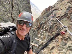 Selfie of man wearing helmet, sunglasses and large backpack clipped into safety gear climbing a small ladder up a rocky mountain
