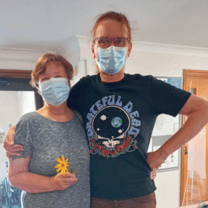 Woman and man wearing blue PPE facemasks. The woman wears a grey top and is holding a yellow flower, the man on the right wears a black T-shirt and has his hand on his hip.