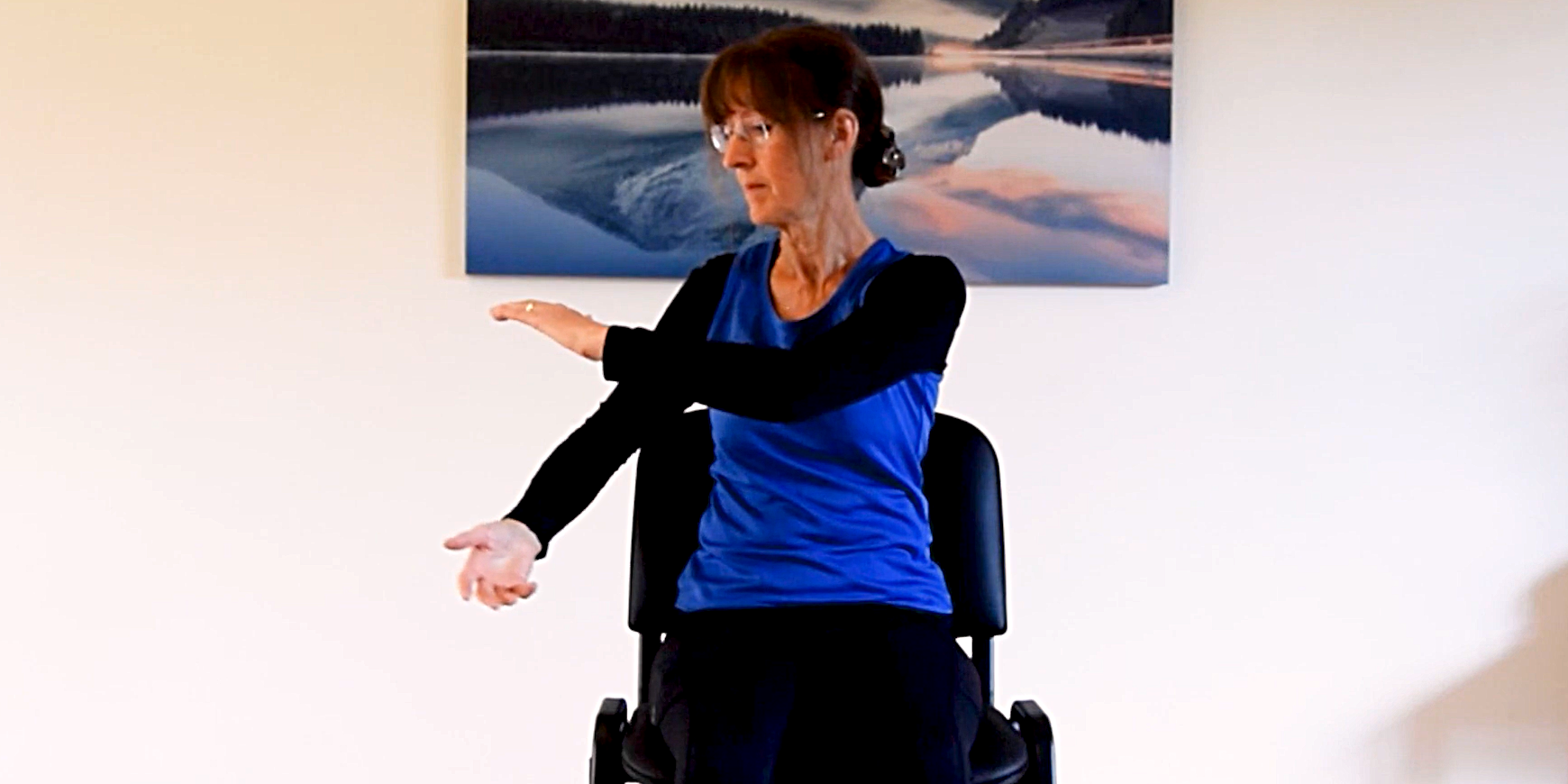 Woman wearing blue top with black sleeves sitting on chair, holding out arms in a tai chi exercise. Behind her are a painting of a lake, and a white wall.