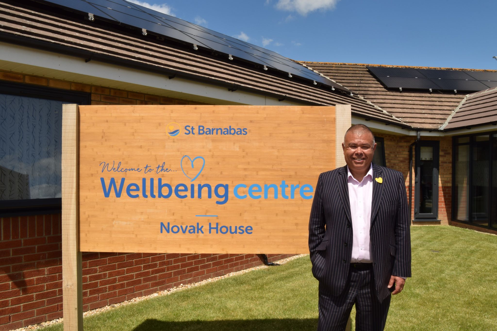 Professor Van Tam, a man in black pinstripe suit with yellow heart pin and light pink shirt, next to wooden sign "Welcome to the Wellbeing Centre - Novak House" with St Barnabas logo. In front of modern building with solar panels