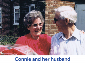 Woman and man with silver hair, the woman wears glasses and a red top, the man on the right a white shirt. White banner underneath and in blue text "Connie and her husband"