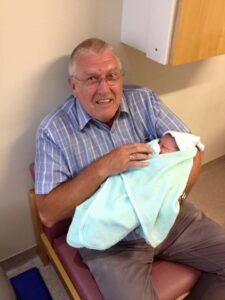 Elderly silver haired man wearing striped blue shirt holding a baby in a light green blanket