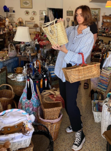 Woman with shoulder length brown hair, blue shirt, holding wicker basket and holding item in charity shop