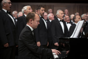 Men singing in a choir wearing black tuxedos. Man in foreground at black piano
