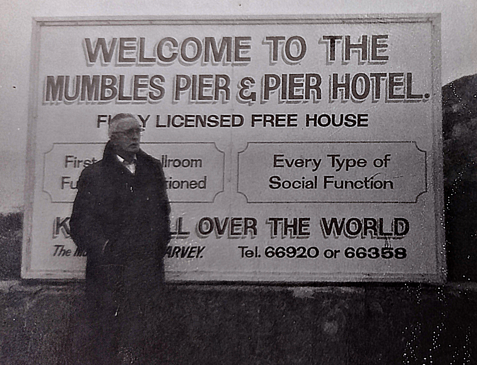 Black and white photo of silver haired man wearing black coat and glasses in front of large sign with text "welcome to the Mumbles Pier and Pier Hotel" with further text about location