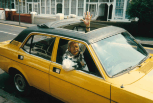 Man with silver hair and moustache in a yellow car with his hand outstretched from the sunroof