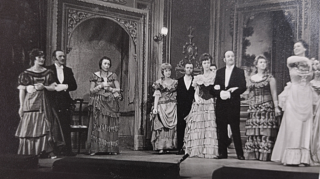 Black and white picture of men and women on stage in a period drama wearing costumes