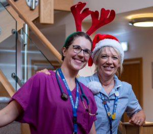 Two women wearing clinical scrubs in purple and blue, and festive reindeer antlers and Christmas hat, standing in stairway