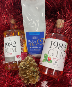 Two bottles of gin and rum along with a white bag of coffee with blue label, on background of red tinsel, with pinecone