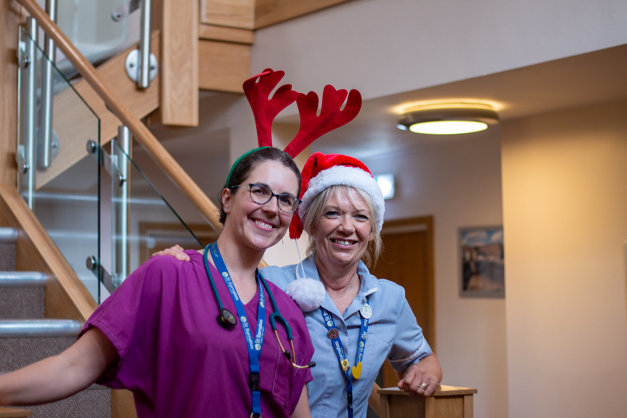 Two women wearing clinical scrubs in purple and blue, and festive reindeer antlers and Christmas hat, standing in stairway