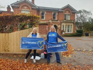 Two women dressed in blue St Barnabas tops holding blue banners with text, in the background is a red brick building - the hospice's Inpatient Unit in Lincoln