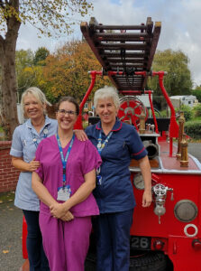 Three women wearing nurse uniforms standing in front of vintage fire engine, smiling and posing