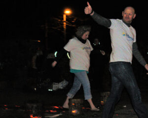 Two people walking over flaming hot coals wearing white tops. Man giving thumbs up