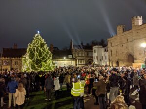 Large Christmas tree with lights, surrounded by dozens of people at night time with Lincoln Castle gate wall in the background