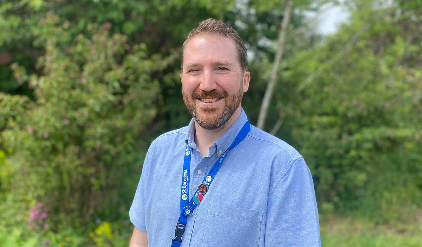 Man with short brown hair and beard wearing blue shirt and dark blue lanyard smiling in outside setting with greenery