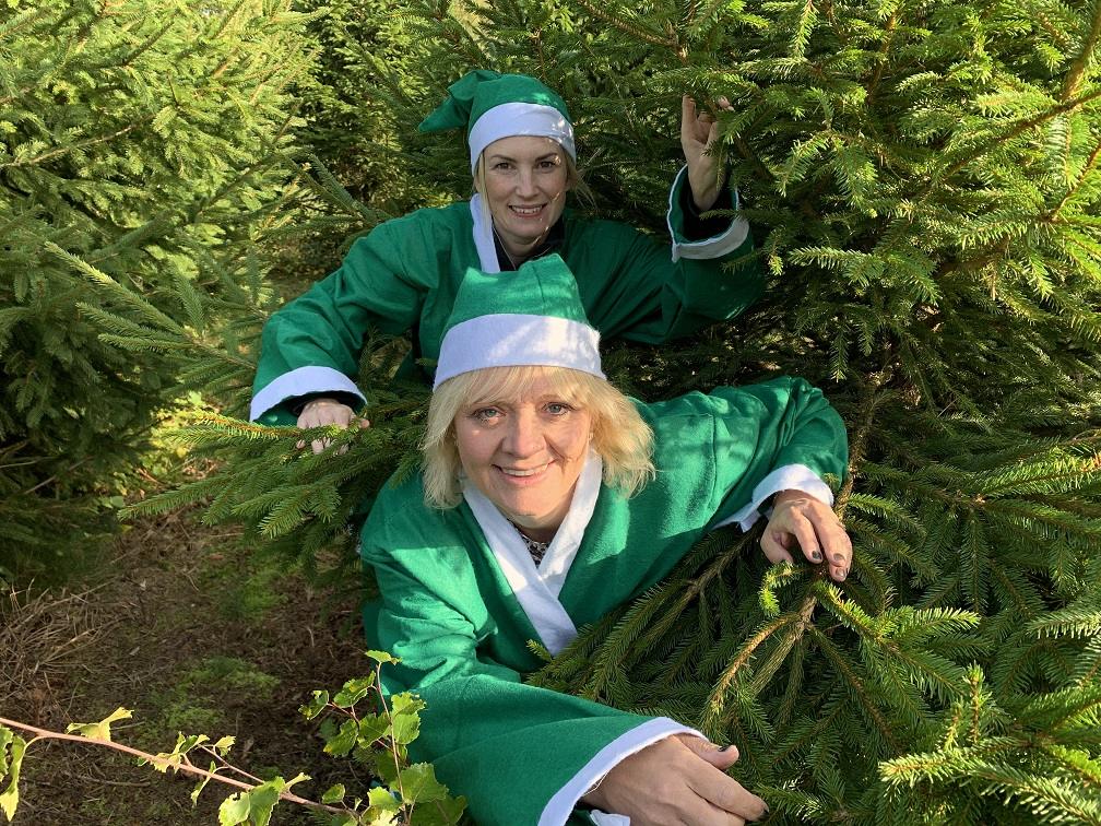 Two blonde women wearing green Christmas elf outfits standing with Christmas trees