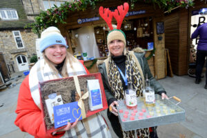 Two women holding items in front of wooden Christmas cabin. Left woman wears blue hat and holds a hamper, woman on the right wears red reindeer antlers and holds a tray with two bottles