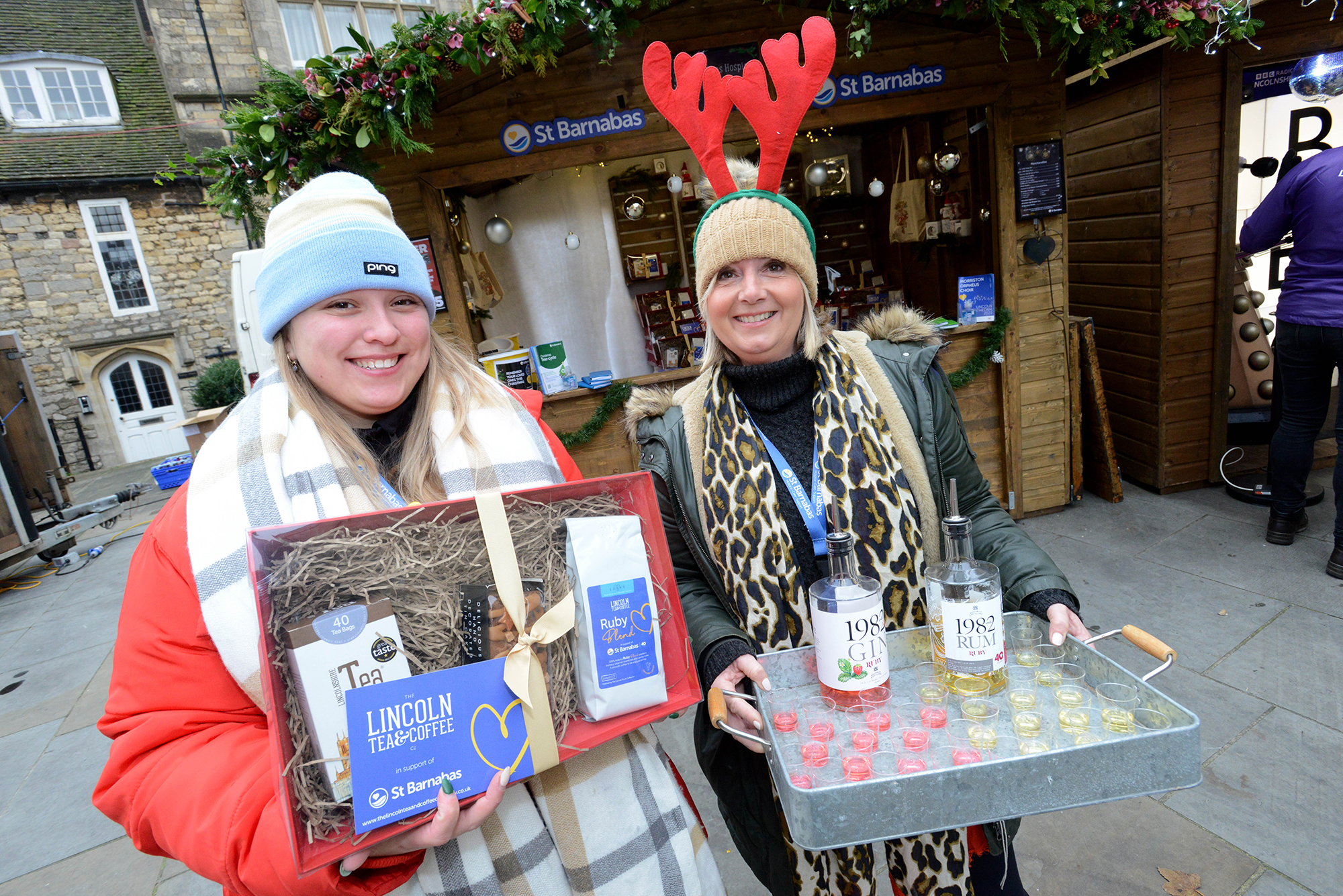 Two women holding items in front of wooden Christmas cabin. Left woman wears blue hat and holds a hamper, woman on the right wears red reindeer antlers and holds a tray with two bottles