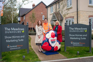 Two women and a man in orange high visibility jacket with large heart sculpture in red, blue and white by housing development with grey signs