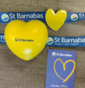 Yellow heart with St Barnabas logo in middle, blue lanyard with yellow pin badge and blue paper with yellow heart