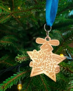 Wooden star-shaped decoration with Light up a Life engraving, on blue ribbon with Christmas tree in background