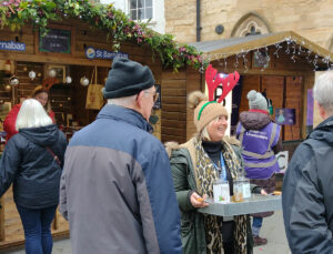 Woman wearing red antlers holding a tray with bottles, talking to people outside at a Christmas MArket