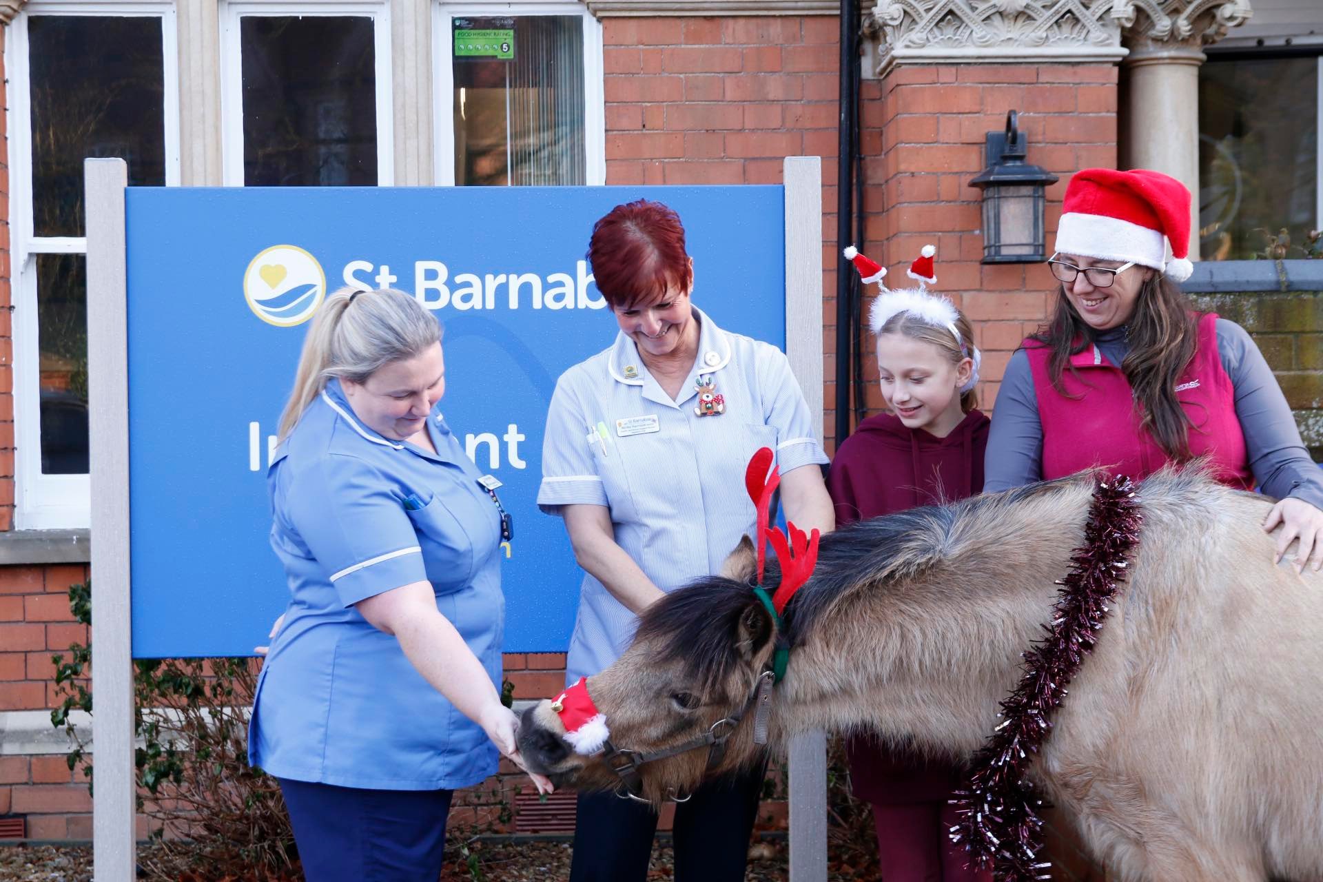 Three women and young girl with brown pony that is wearing red antlers and red nose, in front of blue Inpatient Unit sign